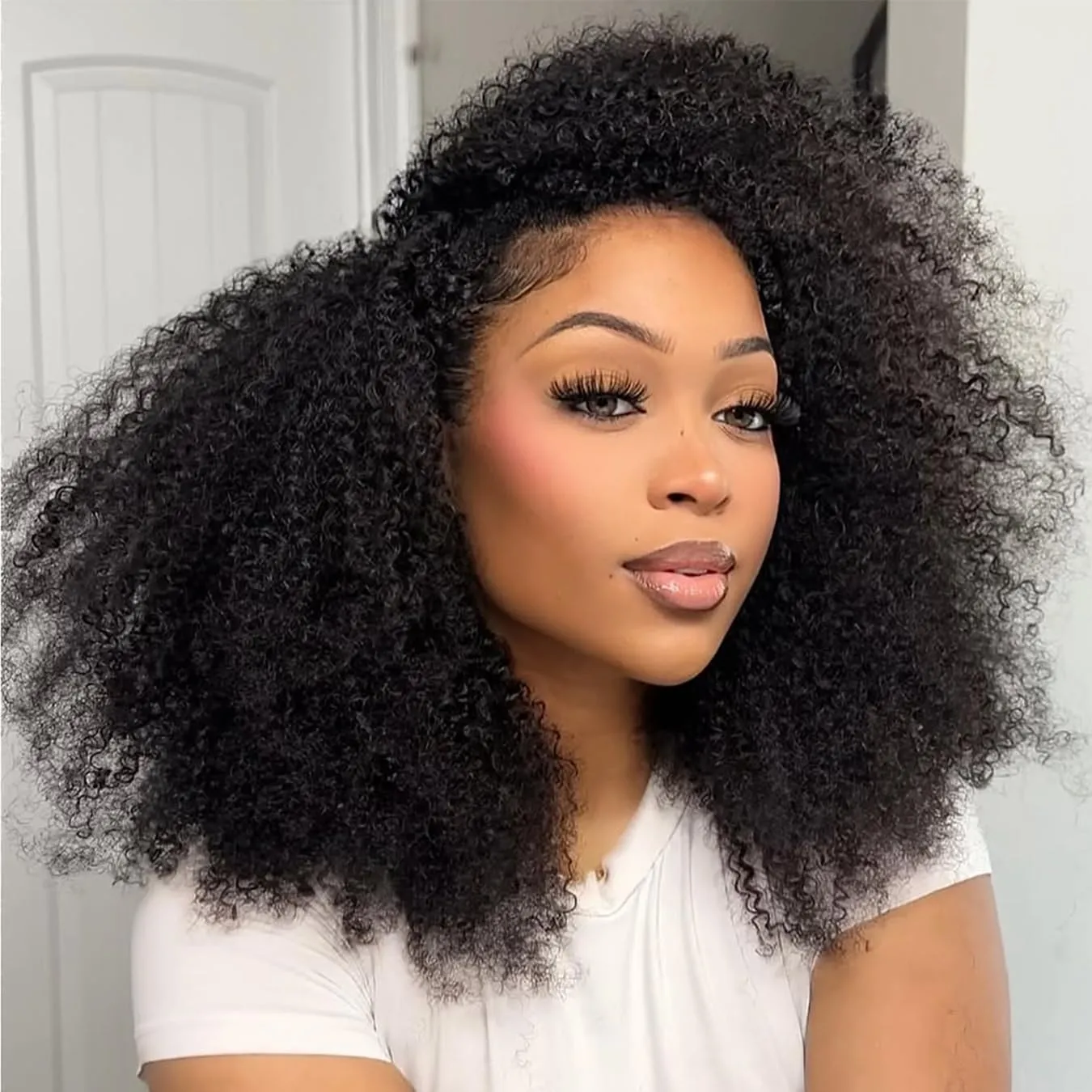 Close-up portrait of a woman with thick, voluminous curly black hair, wearing a white top and soft makeup, looking at the camera.